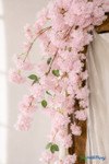41" Beauty Pink Cascading "Spring Blossom" Spray – Extra-Full Silk Floral Waterfall from ShopWildThings.com shown in a close-up wedding arch detail shot with soft pink silk blossoms, green leaves, brown stems, and draped white fabric.