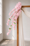41" Beauty Pink Cascading "Spring Blossom" Spray – Extra-Full Silk Floral Waterfall from ShopWildThings.com styled on a simple wooden wedding arch with white draping, natural window light, and cascading pink floral branches for ceremony decor.