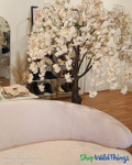 Boutique interior display featuring the ShopWildThings 6-foot Adalee Flowering Cherry Blossom Tree in blush pink, styled in a retail setting with soft neutral tones and gold accents. This artificial tree provides a romantic focal point near the checkout area and is fully compatible with the ShopWildThings Interchangeable Replacement Branch system, allowing boutique owners to refresh their look seasonally with ease.