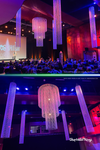 Diamond Crystal Beaded Columns and oversized crystal beaded chandelier suspended over stage at live event, illuminated in purple and blue lighting, creating dramatic ceiling décor for corporate event venue.