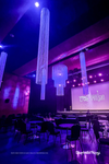 Diamond Crystal Beaded Columns and oversized crystal beaded chandelier suspended over stage at live event, illuminated in purple and blue lighting, creating dramatic ceiling décor for corporate event venue.