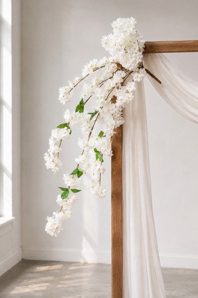 41" White Cascading "Spring Blossom" Spray – Extra-Full Silk Floral Waterfall from ShopWildThings.com attached to a simple wooden wedding arch in natural window light, with white fabric draping and white floral stems cascading downward.