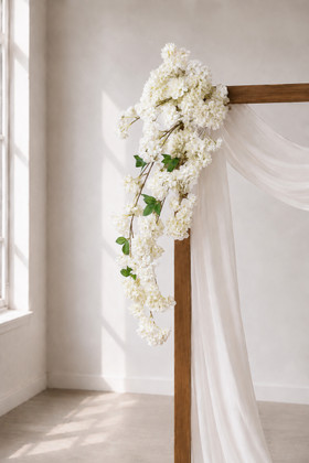 41" Cream White Cascading "Spring Blossom" Spray – Extra-Full Silk Floral Waterfall from ShopWildThings.com attached to a simple wooden wedding arch in natural window light, with white fabric draping and cream white floral stems cascading downward.