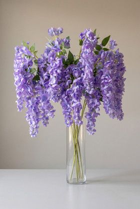 Lavender artificial wisteria floral sprays arranged in a tall clear glass vase with cascading purple blooms and green leaves from ShopWildThings.com