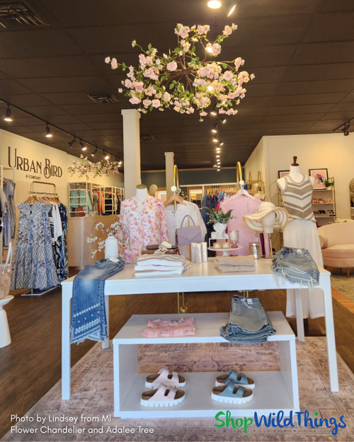 Retail boutique space showcasing the ShopWildThings Roses & Greenery Hanging Canopy, a round floral chandelier measuring 46 inches wide by 16 inches long, suspended over a display table with folded apparel and accessories. The chandelier features artificial roses and greenery in soft pink tones, adding a dramatic, elegant element to the boutique’s visual merchandising strategy.