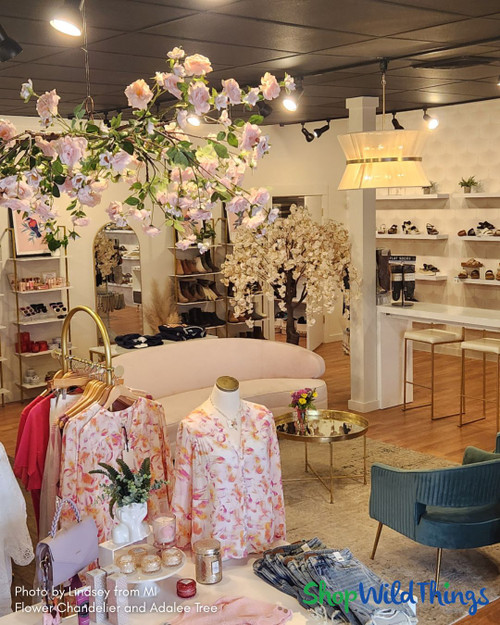 Chic women’s boutique decorated with the ShopWildThings Adalee Cherry Blossom Tree in blush pink and a round floral chandelier canopy of artificial roses and greenery. The tree’s full bloom branches create a romantic atmosphere near a soft blush velvet couch, while the floral chandelier provides overhead interest above a curated display of spring apparel, accessories, and gift items. Ideal for boutique decor and seasonal visual merchandising.