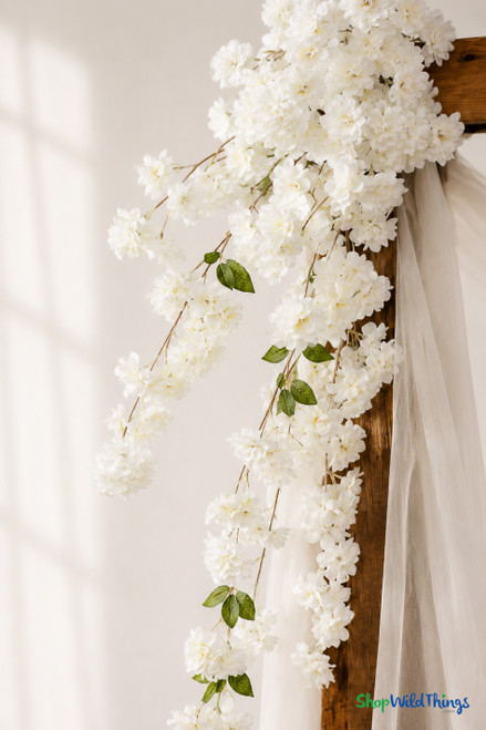 41" Cream White Cascading "Spring Blossom" Spray – Extra-Full Silk Floral Waterfall from ShopWildThings.com styled on a wooden wedding arch with soft white draping, shown in a close-up detail view with cream white blooms and trailing floral sprays.