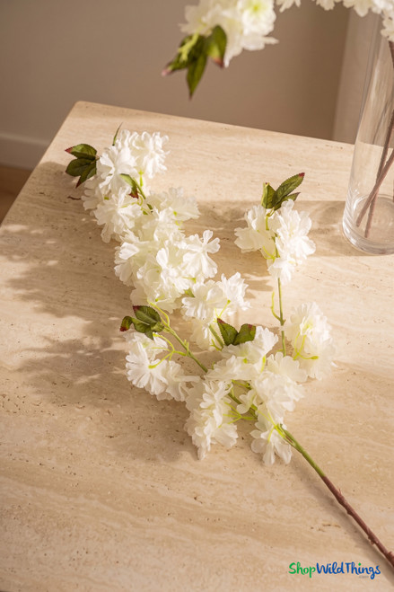Ivory artificial cherry blossom branch detail on travertine tabletop with clear glass vase, ShopWildThings.com silk floral decor