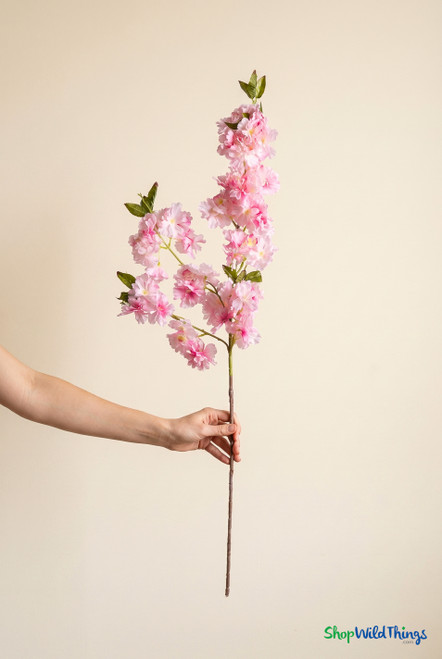 Female hand holding light pink artificial cherry blossom branch for scale against plain wall, ShopWildThings.com silk flower stem