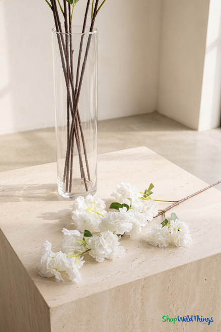 White artificial cherry blossom branch laid beside a clear glass vase on a stone pedestal, showing realistic flowers, leaves, and long brown stems from ShopWildThings.com