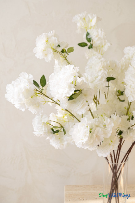 Close-up of artificial white cherry blossom branches with fluffy layered blooms, green leaves, and brown stems in a clear cylinder vase from ShopWildThings.com