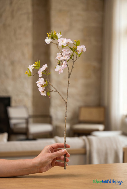 Hand holding artificial cherry blossom branch with light pink and white blossoms for size reference in softly lit interior setting
