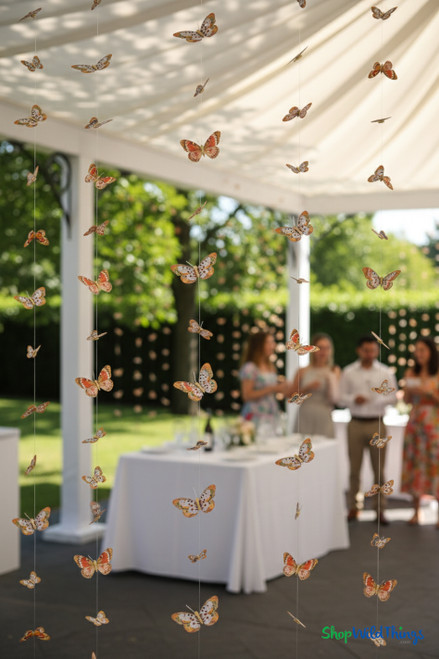 “Madrid” butterfly feather garland strands hanging as whimsical outdoor wedding or garden party backdrop, with floating orange and brown butterflies.