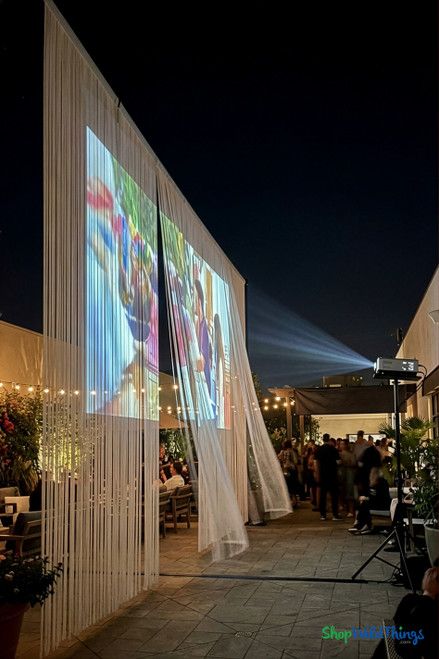 Outdoor evening event with a projected movie displayed on a large white string curtain from ShopWildThings, creating a semi-transparent screen effect; guests gather on a patio with bistro lights and modern seating.