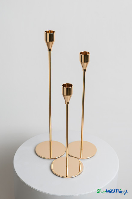 Set of three tall gold metal taper candle holders displayed on white pedestal with elegant varying heights for wedding table decor