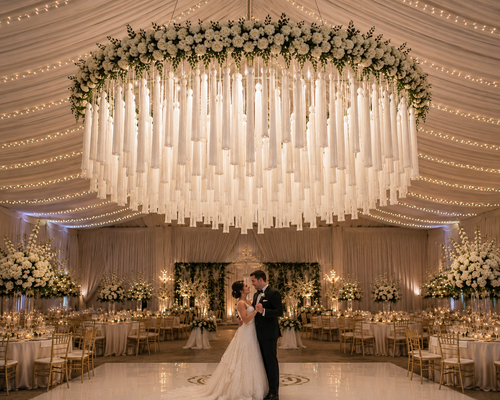 An elegant indoor wedding installation showcasing a custom white tassel chandelier suspended high above the couple’s first dance. The chandelier features layered oversized tassels hanging from a floral-covered circular frame with soft greenery accents. The elevated design creates an airy, luxurious look while allowing the dramatic tassels to frame the dance floor without overwhelming the space. Twinkling ceiling lights and romantic floral arrangements complete the upscale event setting.