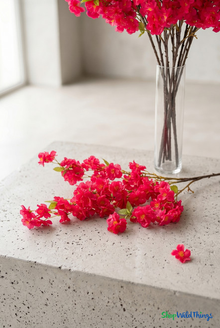 40" Artificial Fuchsia Cherry Blossom Branch – Vivid Spring Flowering Stem for Event Decor from ShopWildThings.com styled with loose bright pink cherry blossom branches on a light stone tabletop beside a clear glass vase