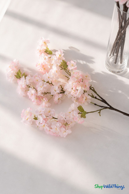 40" Artificial Light Pink Cherry Blossom Branch – Vivid Spring Flowering Stem for Event Decor from ShopWildThings.com styled as a soft window-lit flat lay with pale pink blossoms and clear vase.
