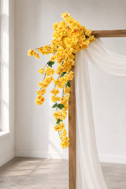 41" Yellow Cascading "Spring Blossom" Spray – Extra-Full Silk Floral Waterfall from ShopWildThings.com attached to a simple wooden wedding arch in natural window light, with white fabric draping and yellow floral stems cascading downward.