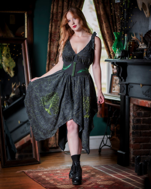 Woman posing in the Morrigan Apron Dress in grey with green embroidery in front of a mirror.