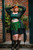 Model posing in front of brick wall in Spellcaster Tailcoat in green, paired with green Elven Blade  Mini Skirt, and Dark Pixie Skirt.