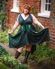 Woman in elf ears and flower crown posing in Morrigan Apron Dress in black and green  in front of brick building, holding out the ends of the dress. The dress is paired with forest green Selene Corset.