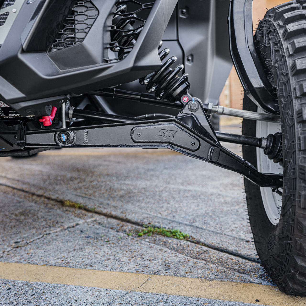 Can-Am Maverick R High Clearance Boxed Lower A-Arms, close-up of the black boxed lower A-arm installed beneath the vehicle’s suspension beside the front wheel. - S3 Power Sports