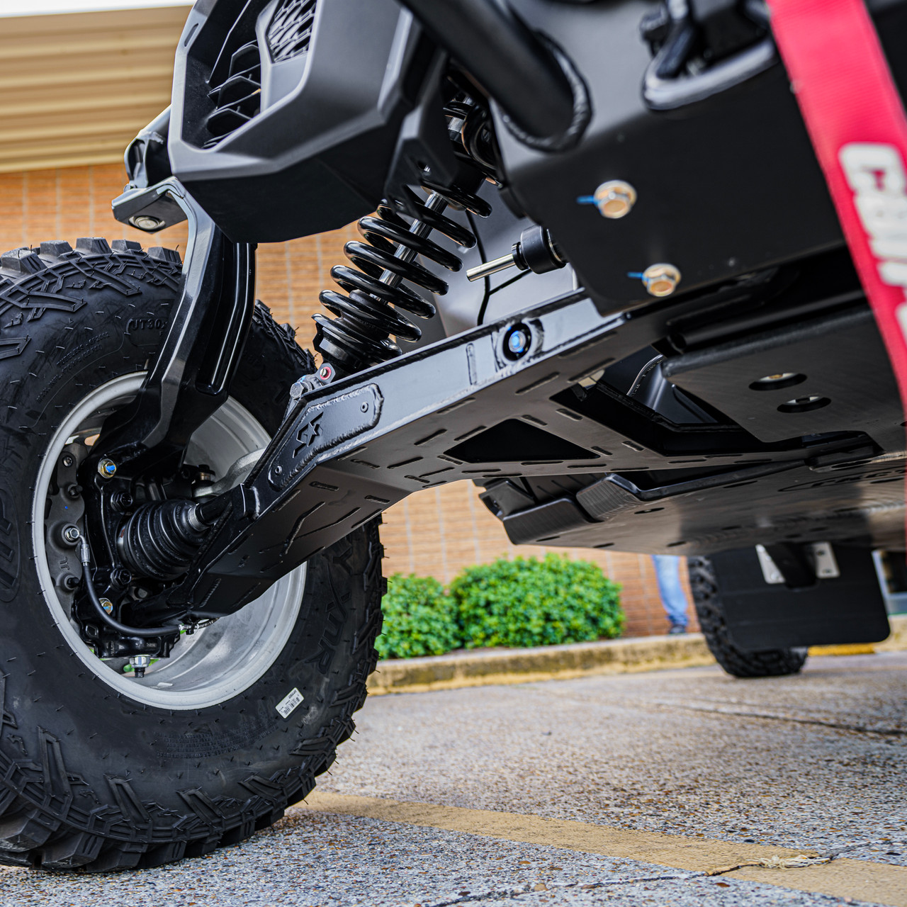 Can-Am Maverick R High Clearance Boxed Lower A-Arms, close-up of the black boxed lower A-arm installed beneath the front suspension and large off-road tire - S3 Power Sports
