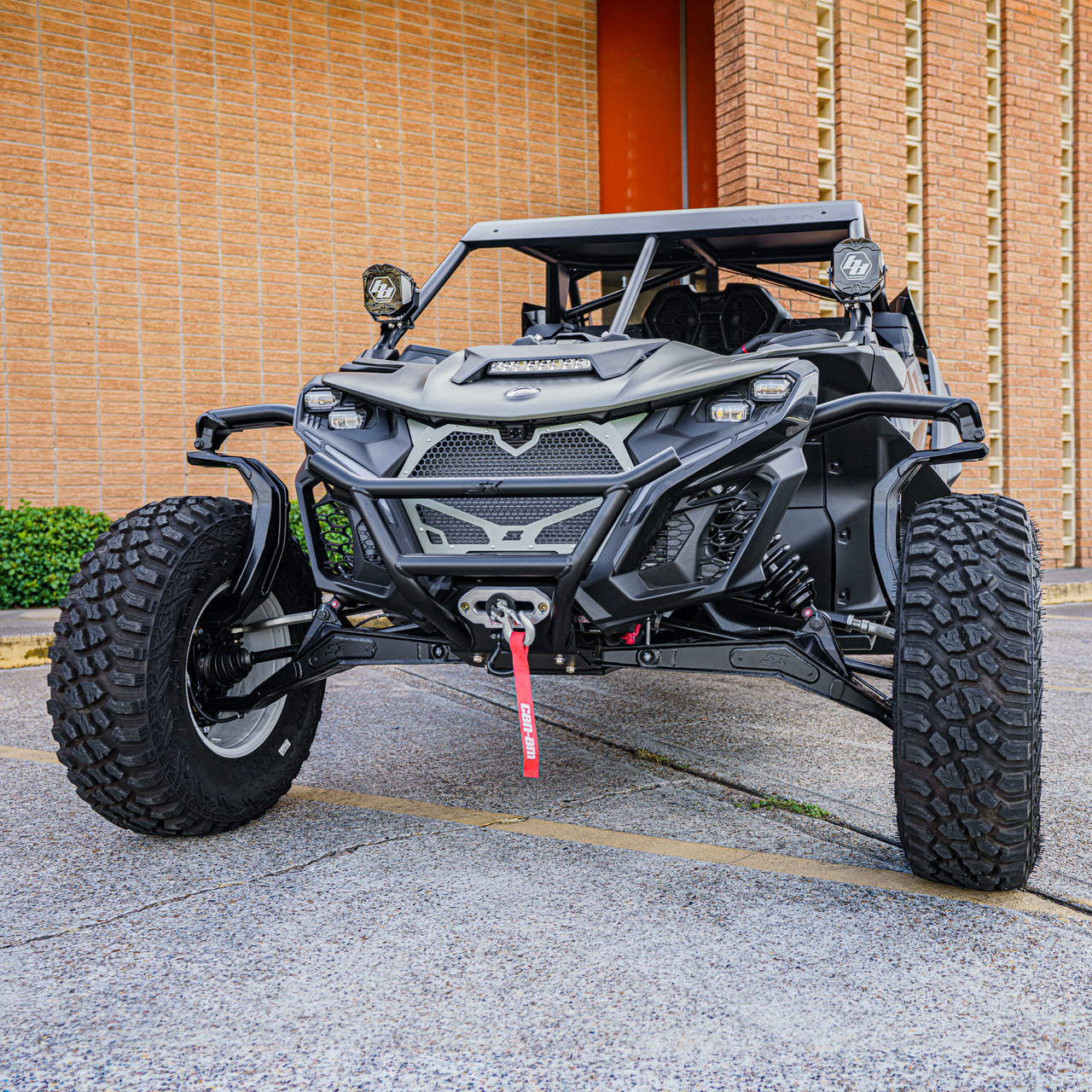 2026 Can-Am Maverick R, green and black side-by-side UTV shown from the front with rugged off-road tires, a roll cage, LED headlights, and a front winch, parked outside a brick building.