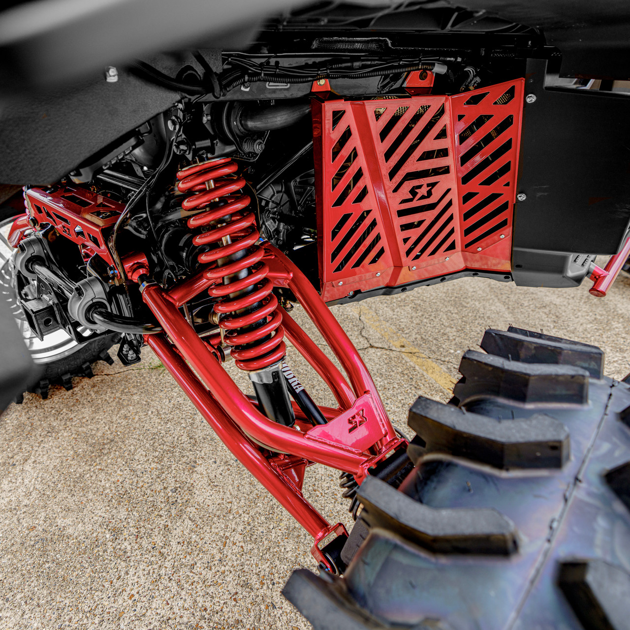 2026 Can-Am Defender HD11, Silver / Red / Black, close-up of red suspension components, coil spring, control arms, and protective panels beside a rugged off-road tire.