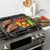 nordic ware dual top grill griddle on stovetop with steaks, pressed potatoes and veggies, grill press on counter in background with cutting board of ingredients