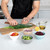 Four white ceramic mini prep bowls with chopped vegetables beside person cutting ingredients on wooden board