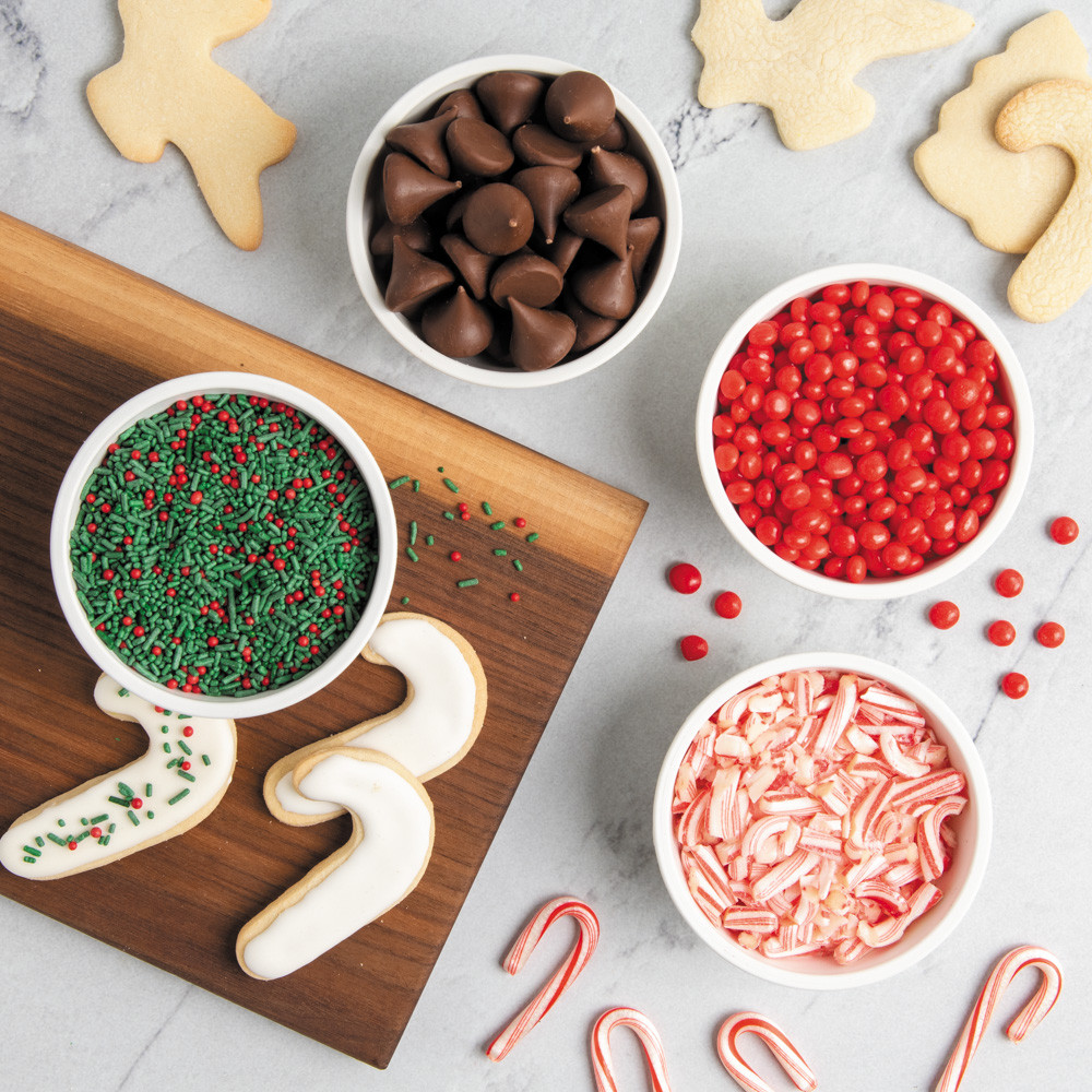 Four white mini bowls arranged on white countertop with crushed candy cane, red and green sprinkles, and red candies 