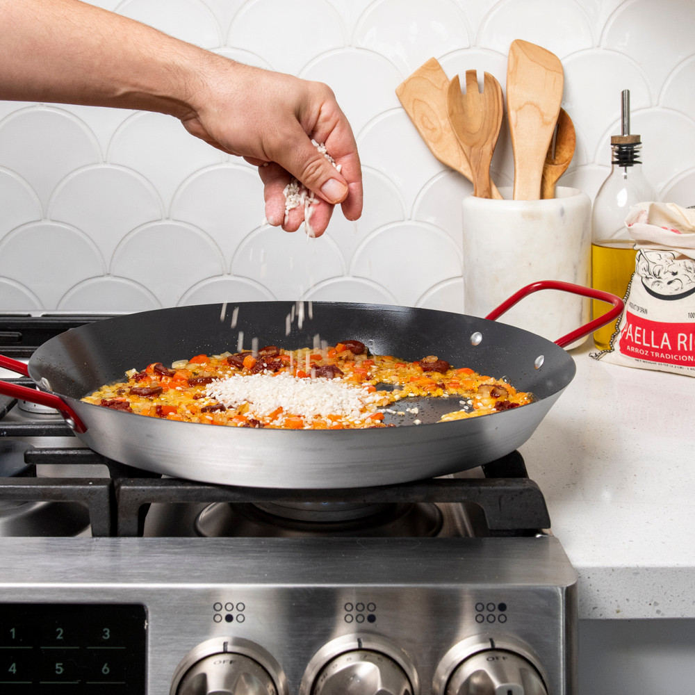 paella pan with hand sprinkling saffron rice, shrimp, mussels, and vegetables arranged on stovetop