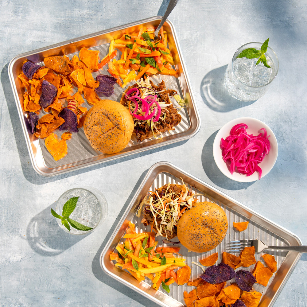 Two Nordic Ware aluminum prep trays holding grilled burgers, golden fries and chips in outdoor BBQ setting