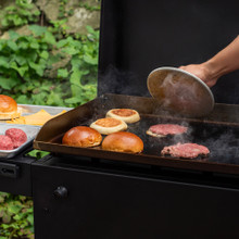 hand holding nordic ware weighted grill press above freshly pressed burger patties on grill with toasting buns