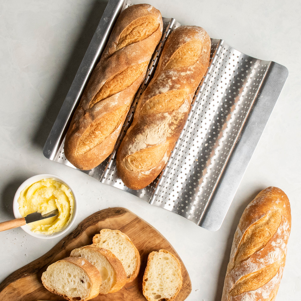 two fresh baked baguettes nested in a well of Nordic Ware baguette & french bread pan; one baguette cut into slices on cutting board