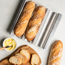 two fresh baked baguettes nested in a well of Nordic Ware baguette & french bread pan; one baguette cut into slices on cutting board