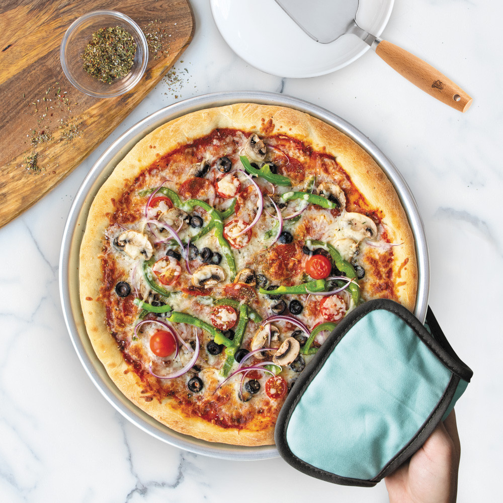 hand with hot pad holding baked pizza in nordic ware deep dish pizza pan, cake server resting on white plate