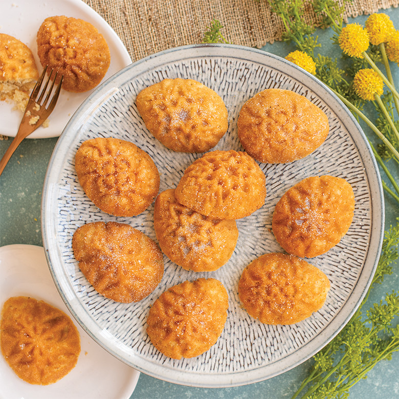 Six golden egg-shaped cakelets with ornate floral designs on white plates, blue background with fresh flowers