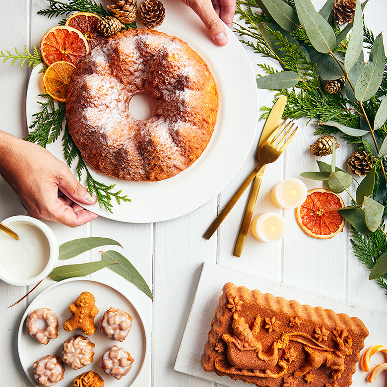 Nordic Ware Holiday Teacakes pan with baked golden mini cakes, pine forest bundt, and Santa sleigh loaf displayed