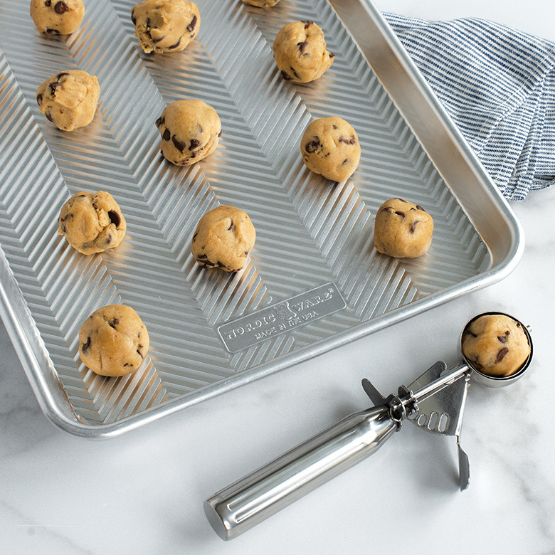 Silver stainless steel cookie scoop holding chocolate chip cookie dough with unbaked dough balls on baking sheet