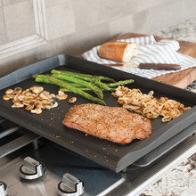 Nordic Ware aluminum double backsplash griddle on stovetop with grilled steak, sautéed onions, mushrooms and asparagus