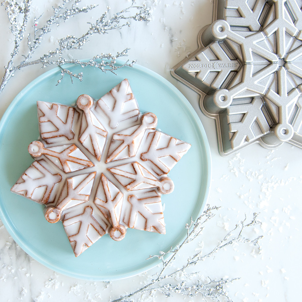 Glazed bundt cake with snowflake design next to Nordic Ware aluminum snowflake baking pan on white surface