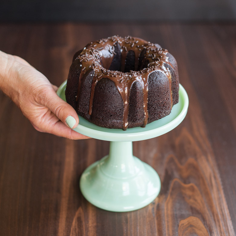 Hand holding small chocolate bundt cake on white pedestal plate, baked in Nordic Ware 3-cup bundt pan