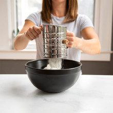 Woman using Nordic Ware aluminum flour sifter with red handle, sifting white flour into glass mixing bowl