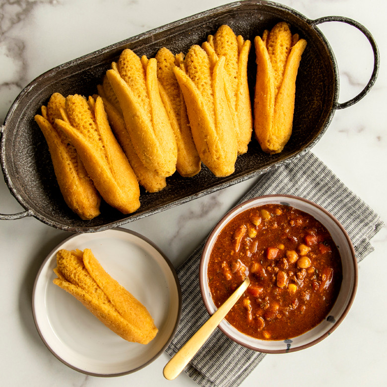 Golden cornbread cakes baked in Nordic Ware corn-shaped baking pan with bowl of chili on marble surface