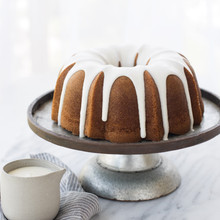 Golden bundt cake with white glaze on silver pedestal stand, small glass bowl of extra glaze nearby