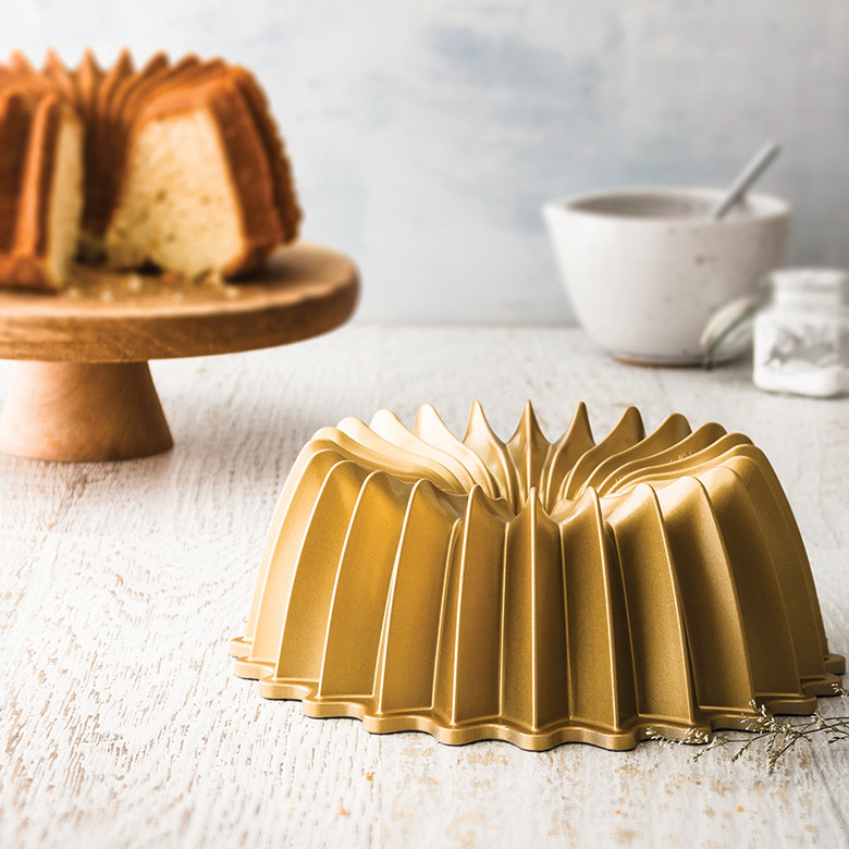 Nordic Ware aluminum Brilliance bundt pan with geometric fluted design, sliced golden bundt cake in background