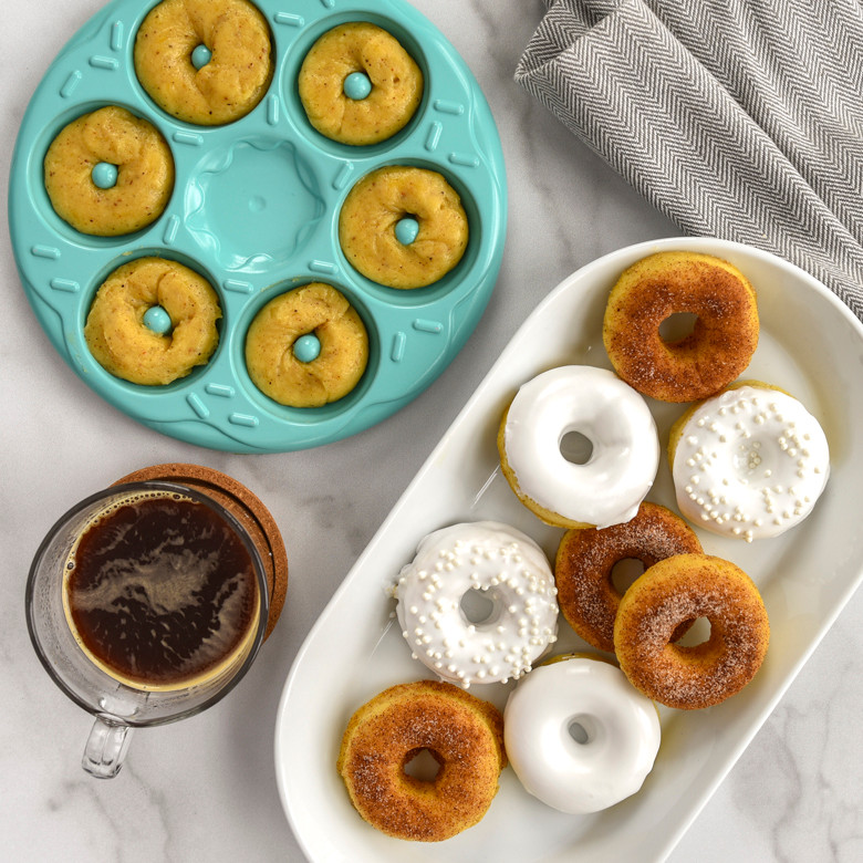 Nordic Ware aluminum donut bites pan with raw batter in cavities, beside plate of glazed mini donuts and coffee cup